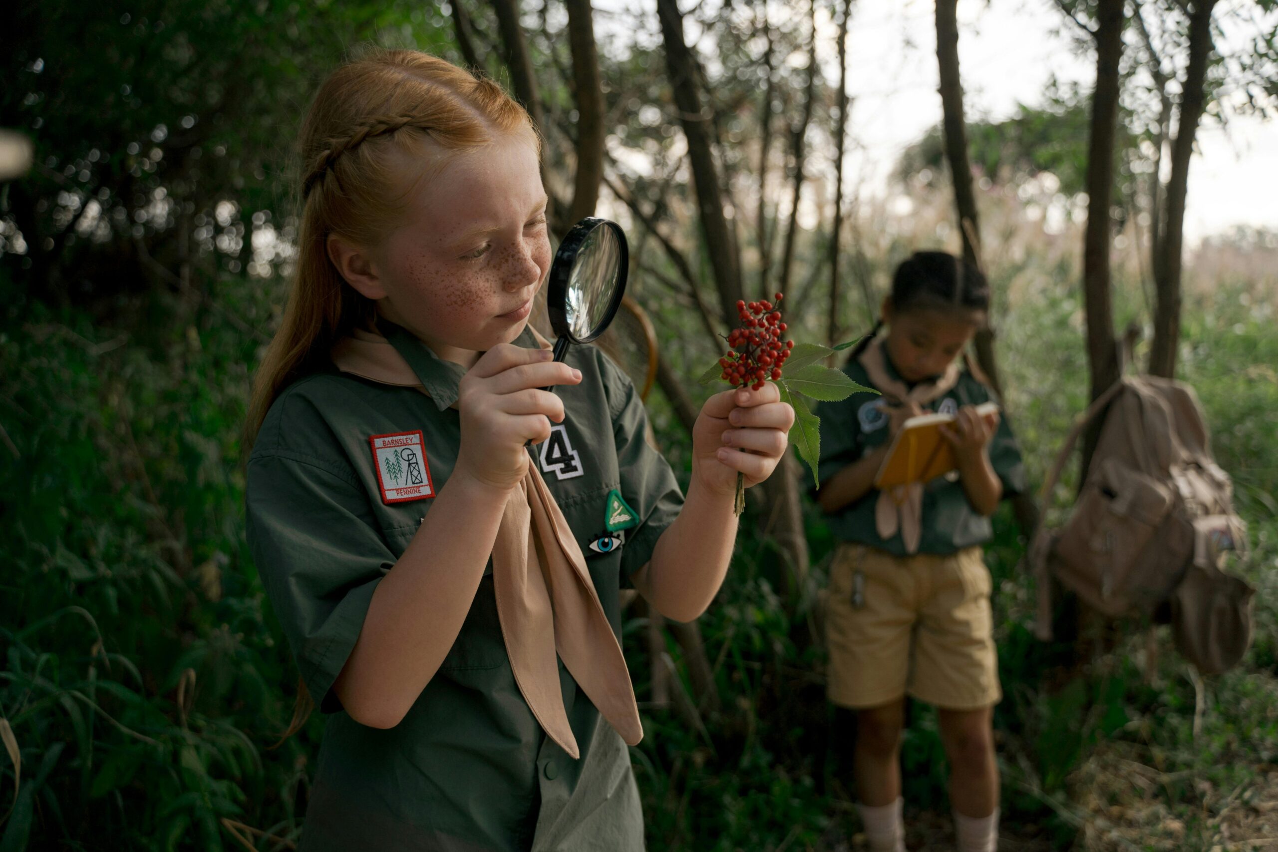 Children exploring nature at a summer scouting camp, using magnifying glasses and taking notes.